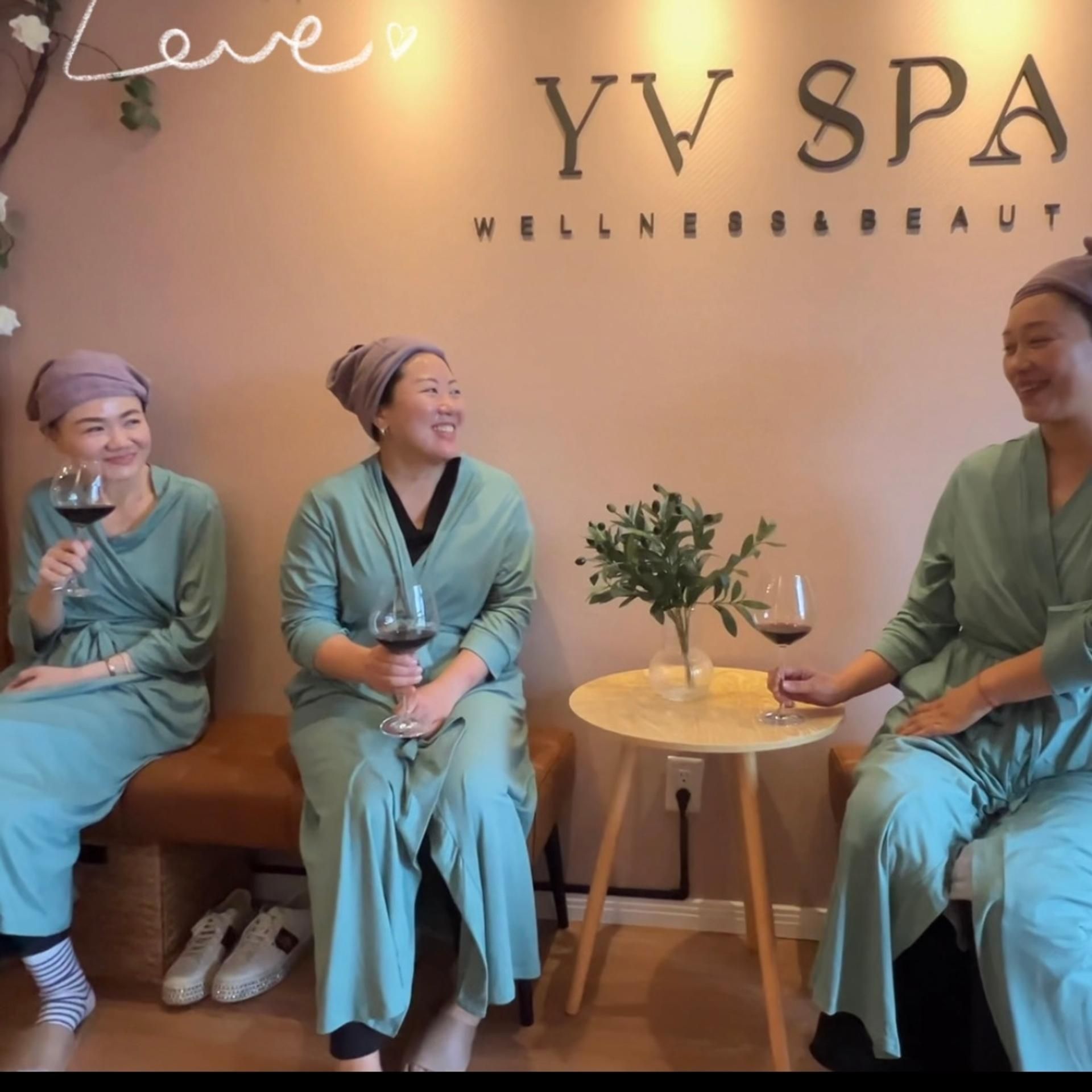 Three women in robes and headscarves, smiling, holding wine glasses, in a spa.