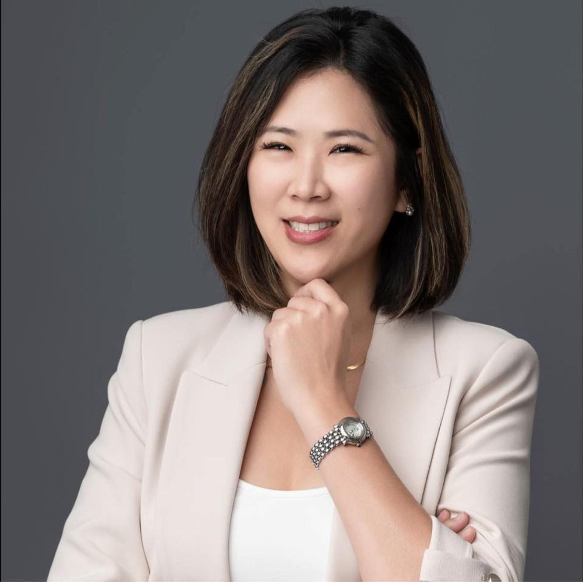 Woman in light blazer, hand on chin, smiling, wearing a watch, neutral background.