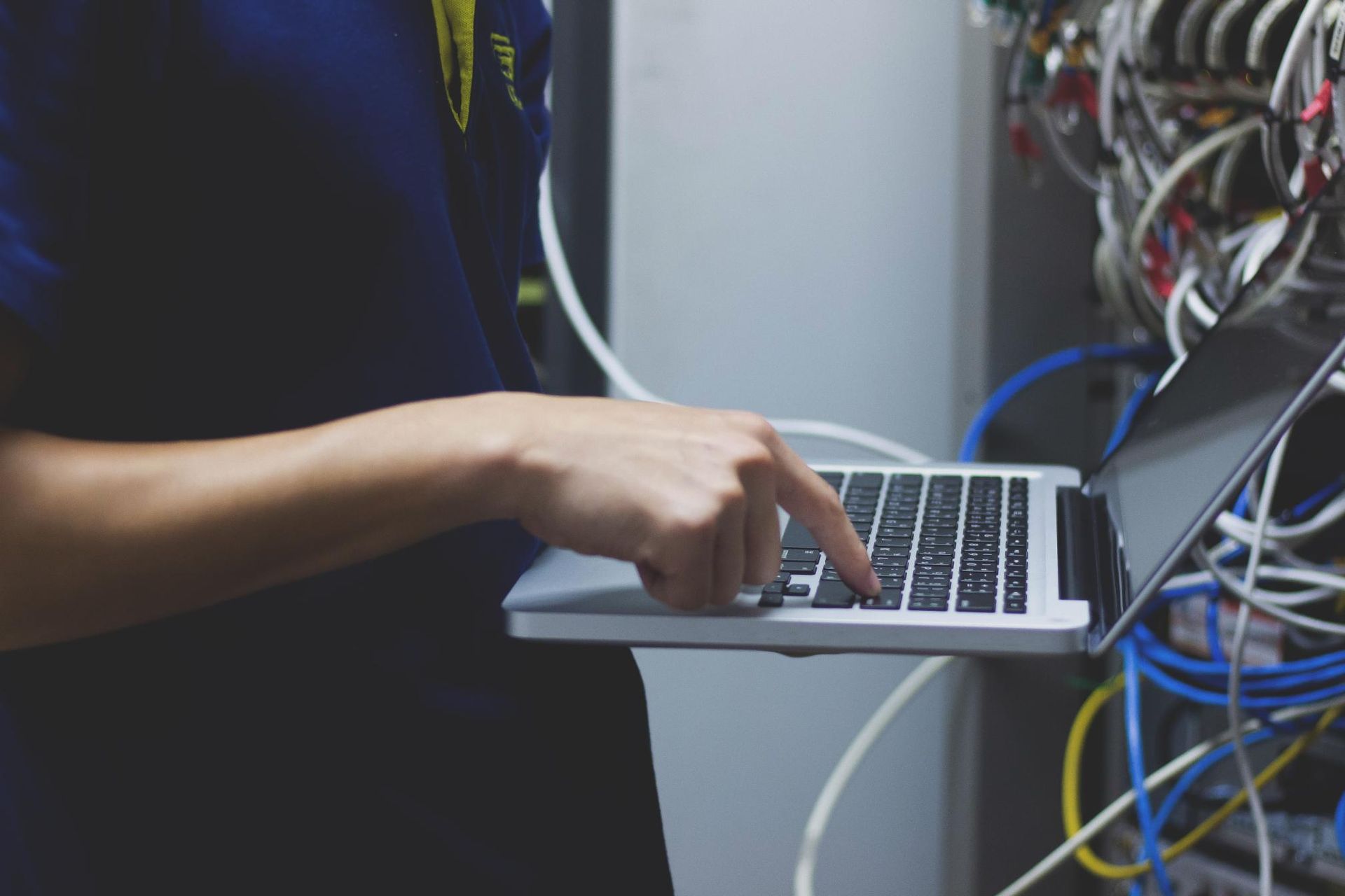 Person using a laptop in a server room, typing with one hand, cables in the background.
