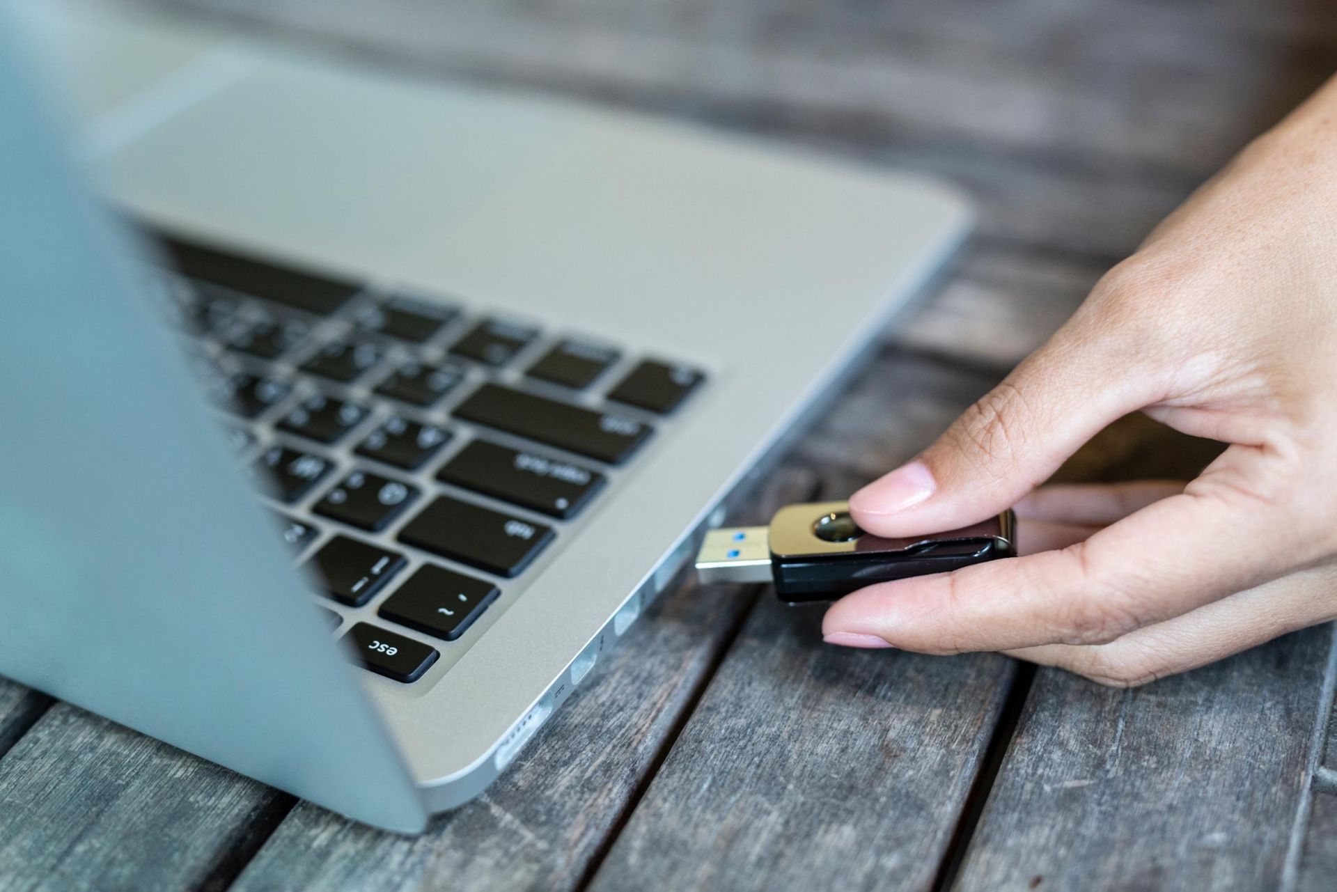 Hand inserting a USB flash drive into a laptop port on a wooden table.