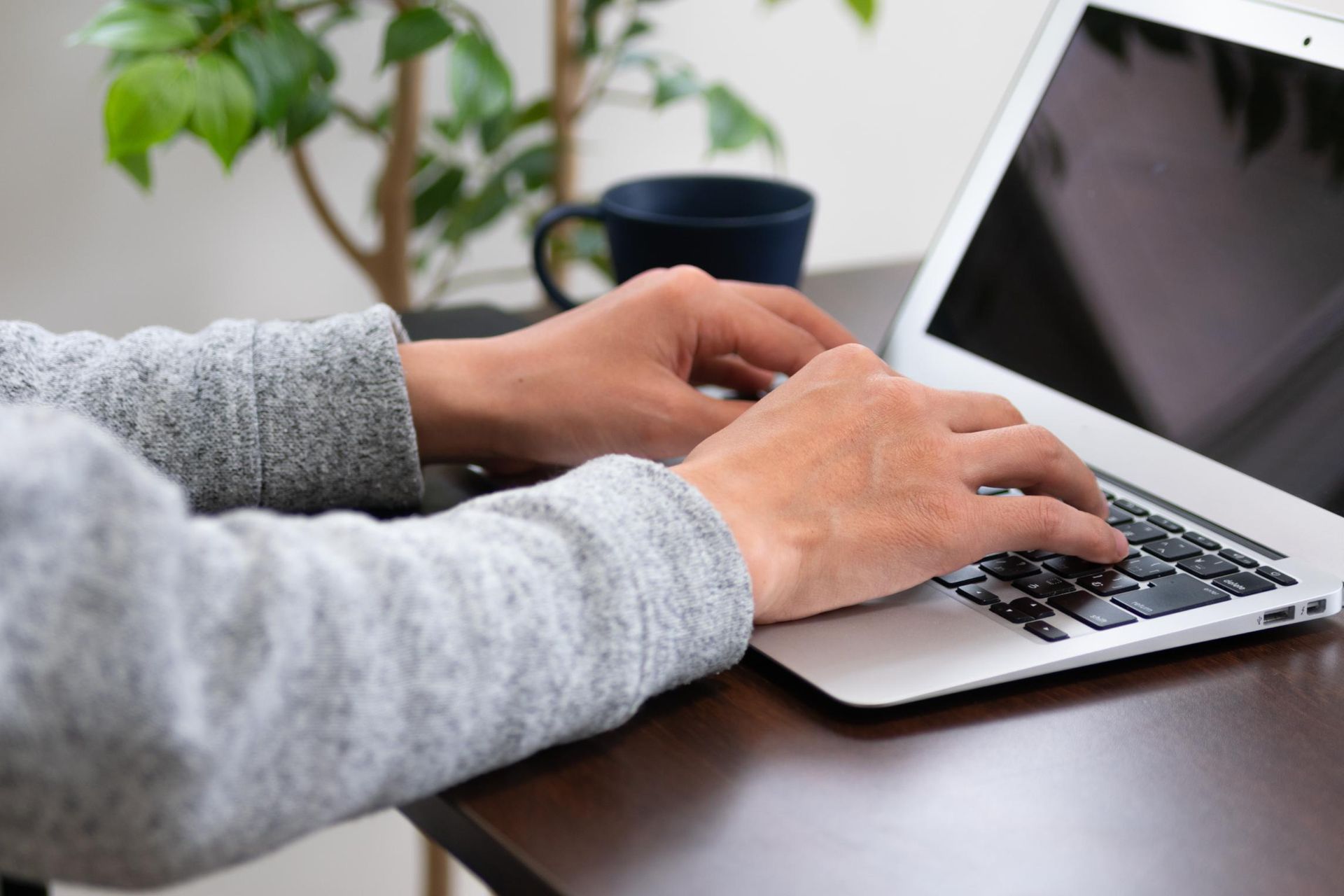 Hands typing on a laptop at a wooden desk, mug and plant in background.