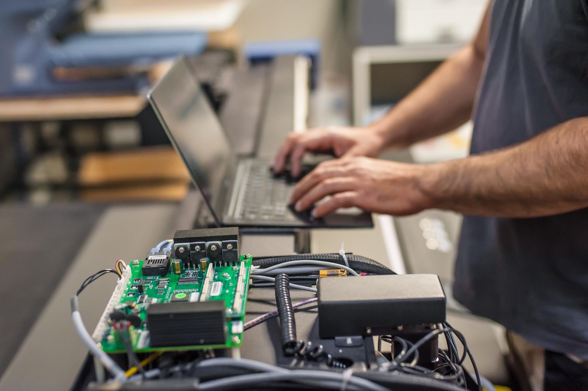 Person using laptop, possibly programming or testing electronic circuit board on a machine in a workshop.
