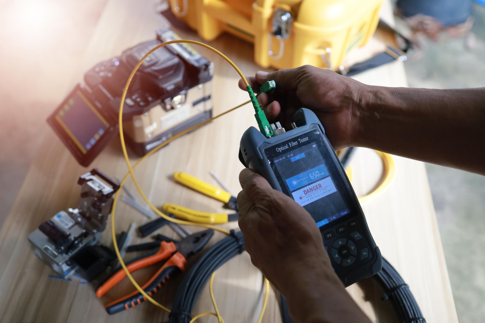 Person using a handheld fiber optic cable tester on a wooden surface, with tools and a yellow case in the background.