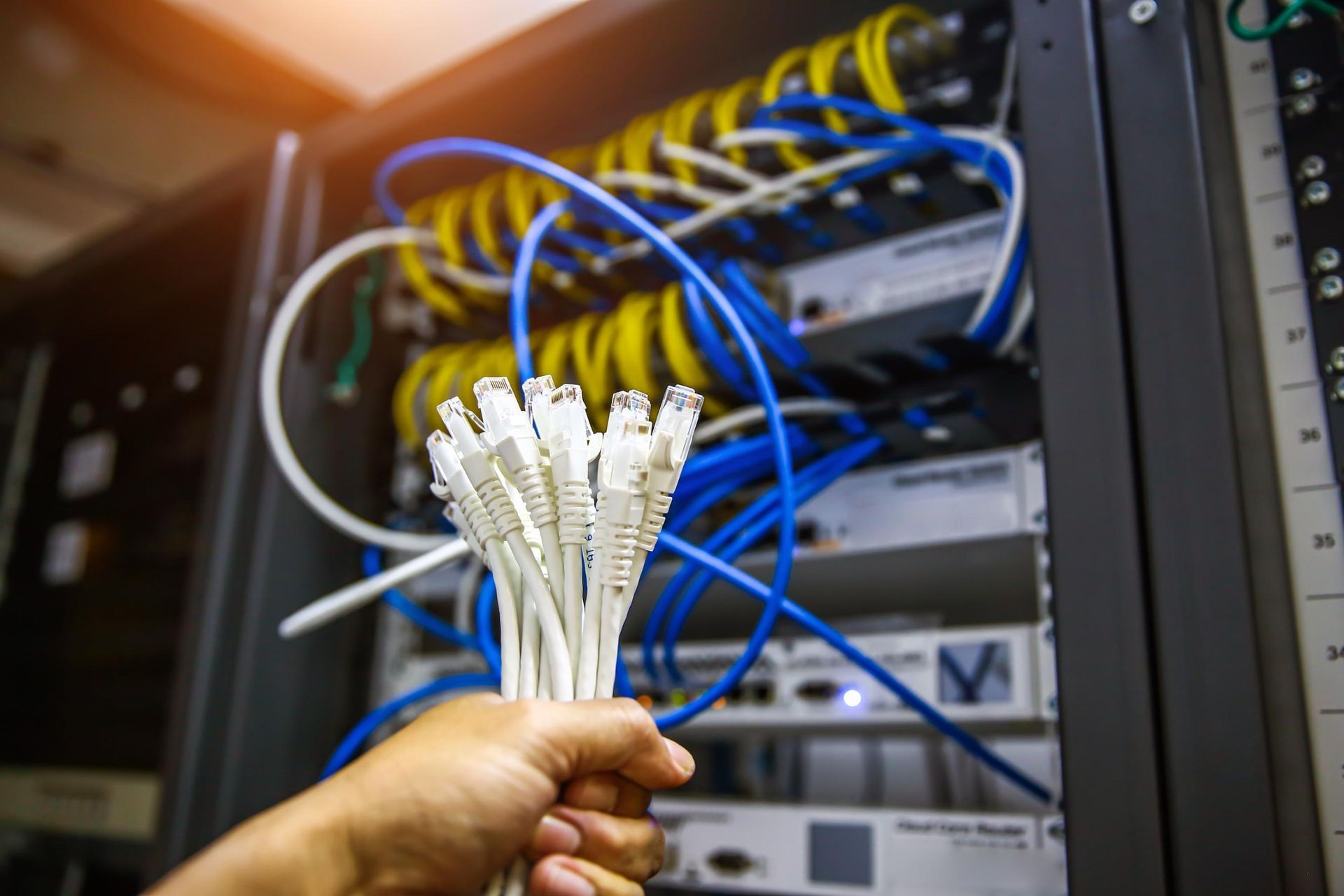 Hand holding a bundle of white Ethernet cables in front of a server rack with blue and yellow cables.