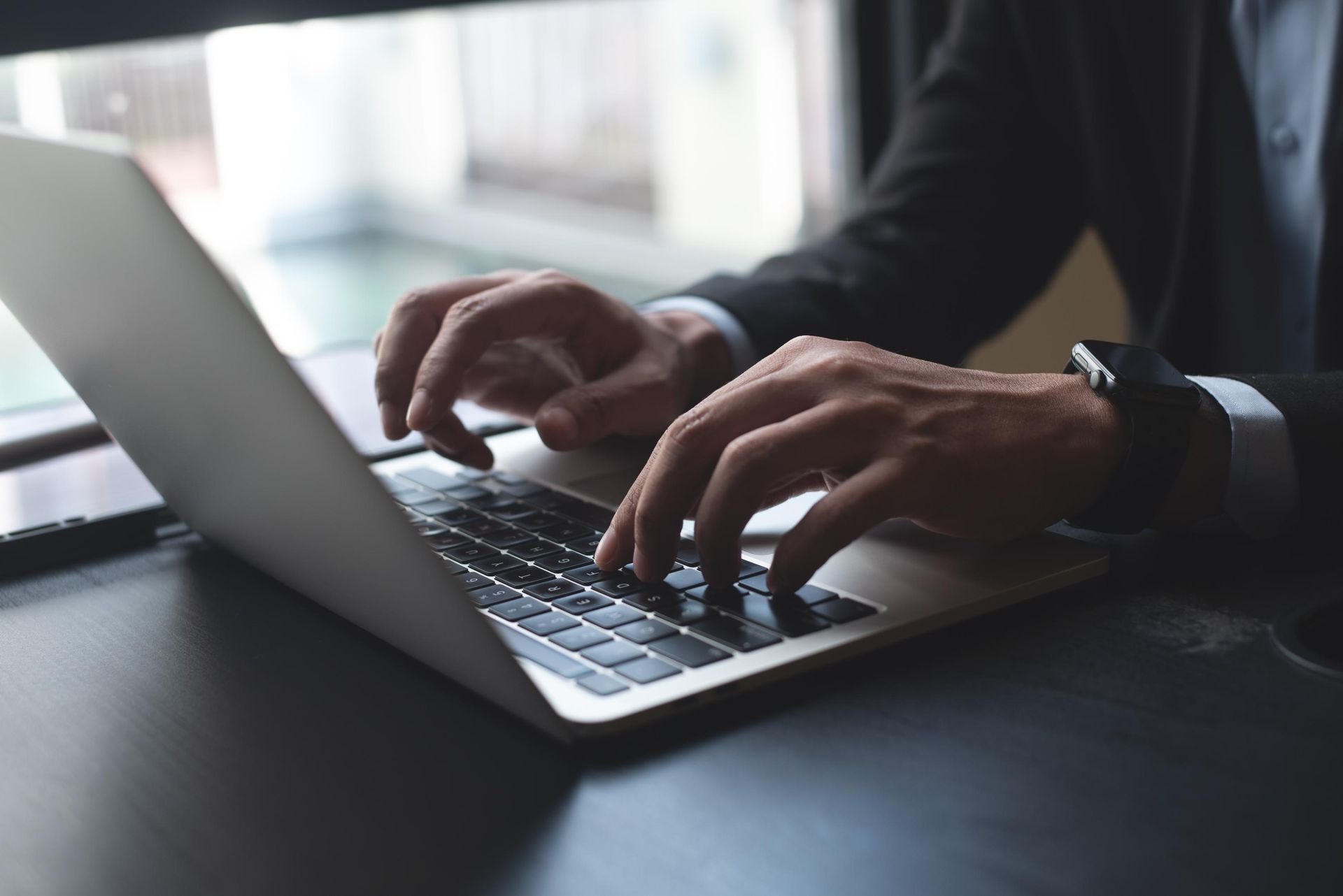 Person in a suit typing on a laptop at a desk with window in the background.