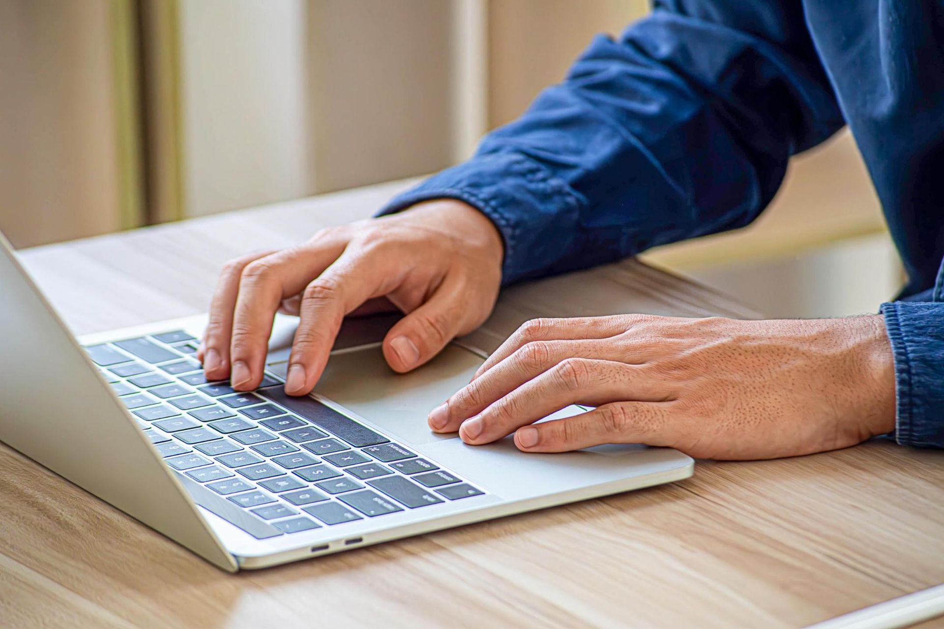 Person typing on a laptop, hands on keyboard, blue shirt, on a wooden desk.