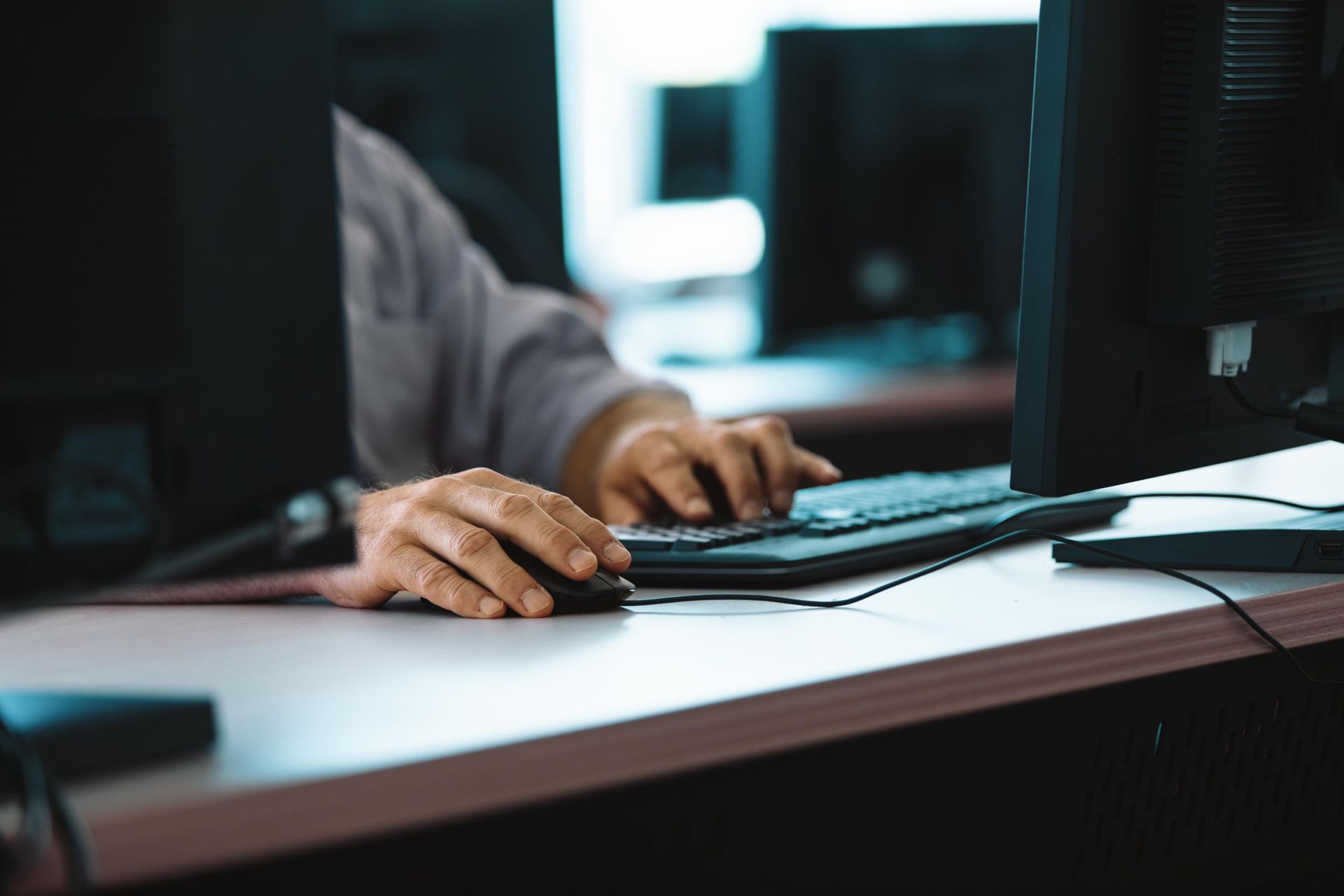 Hands typing on a keyboard, with a mouse on a desk and computer monitors in the background.