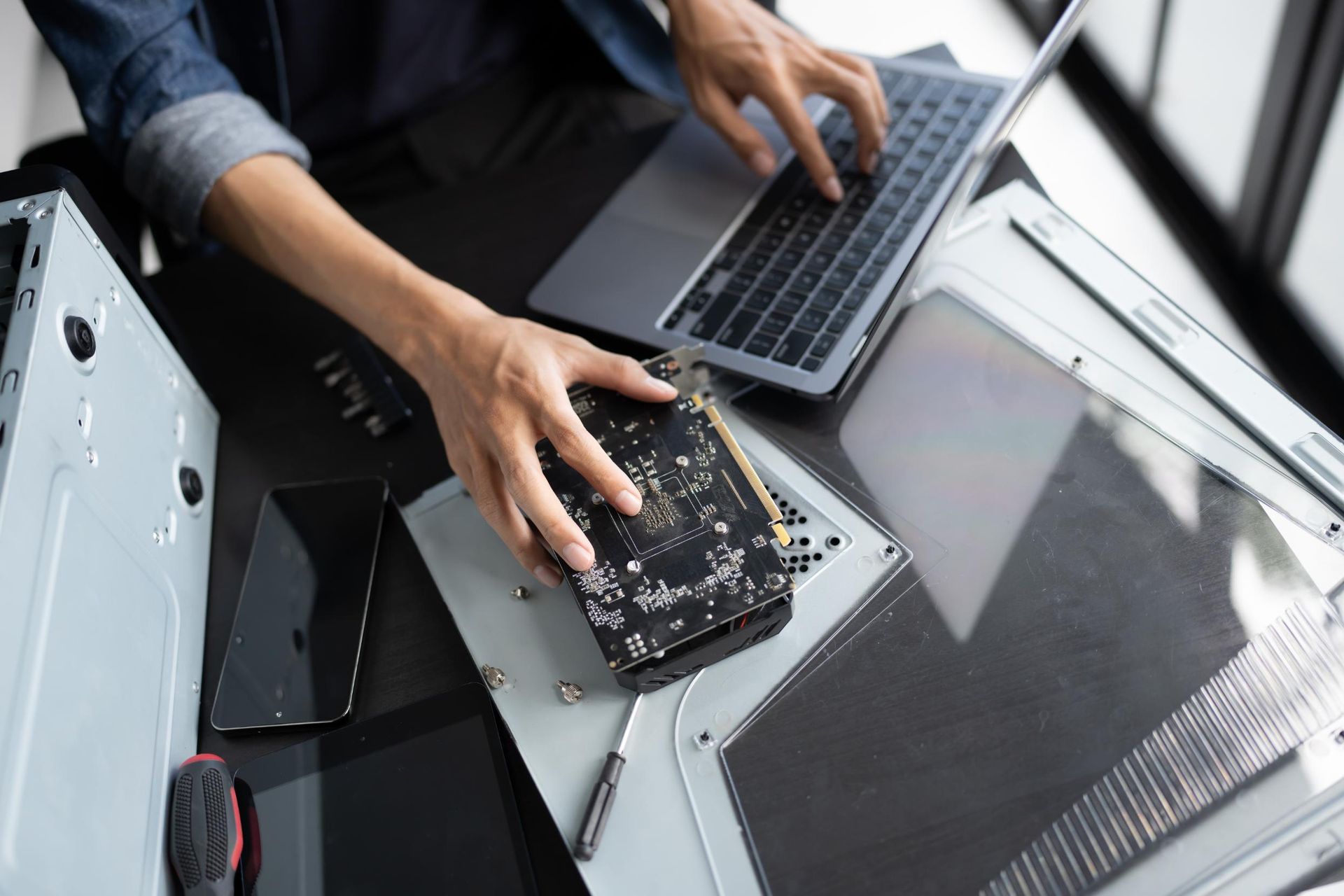 Person repairs computer components at a desk with a laptop, holding a circuit board.