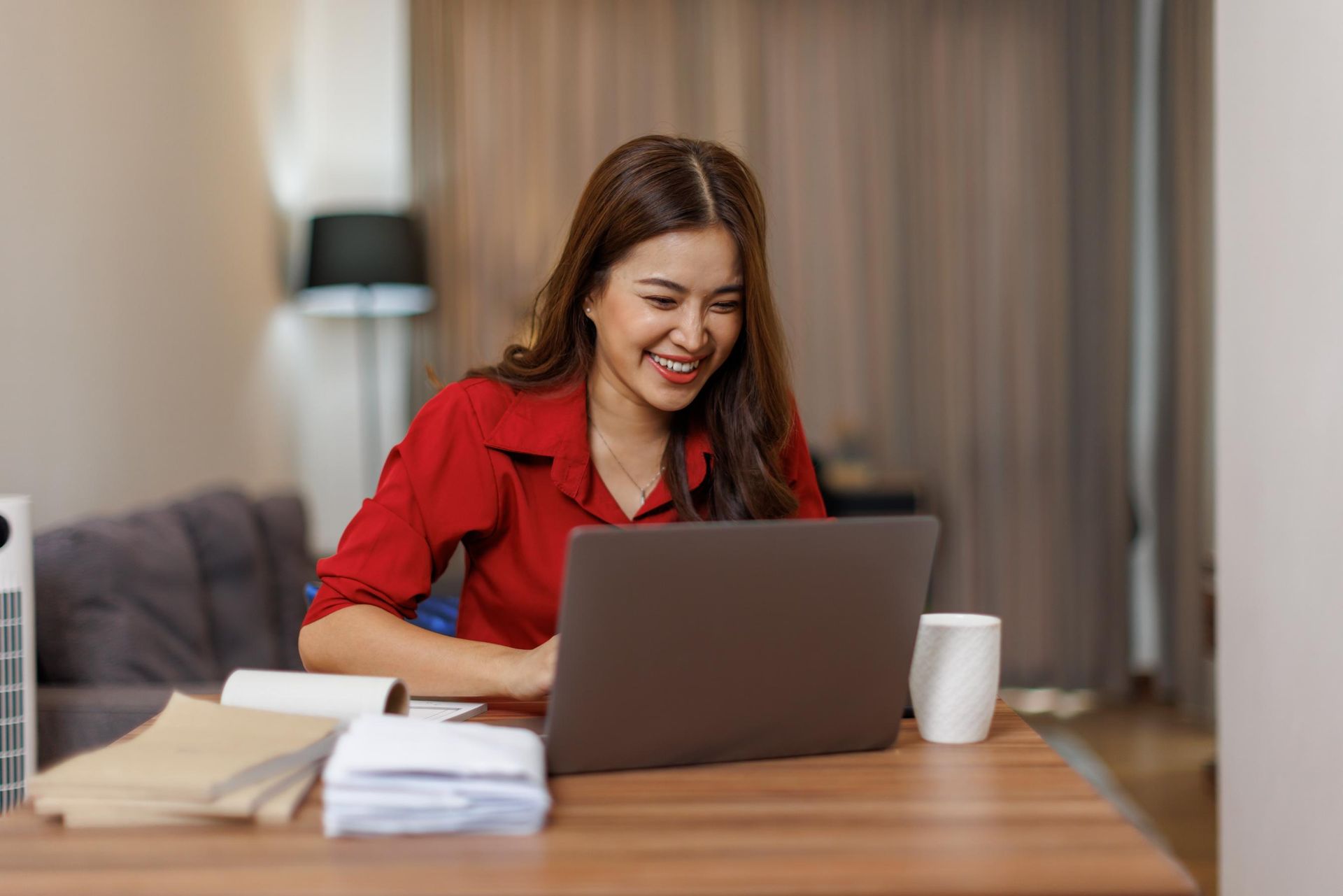 Woman smiles while working on a laptop at a table in a home office setting.