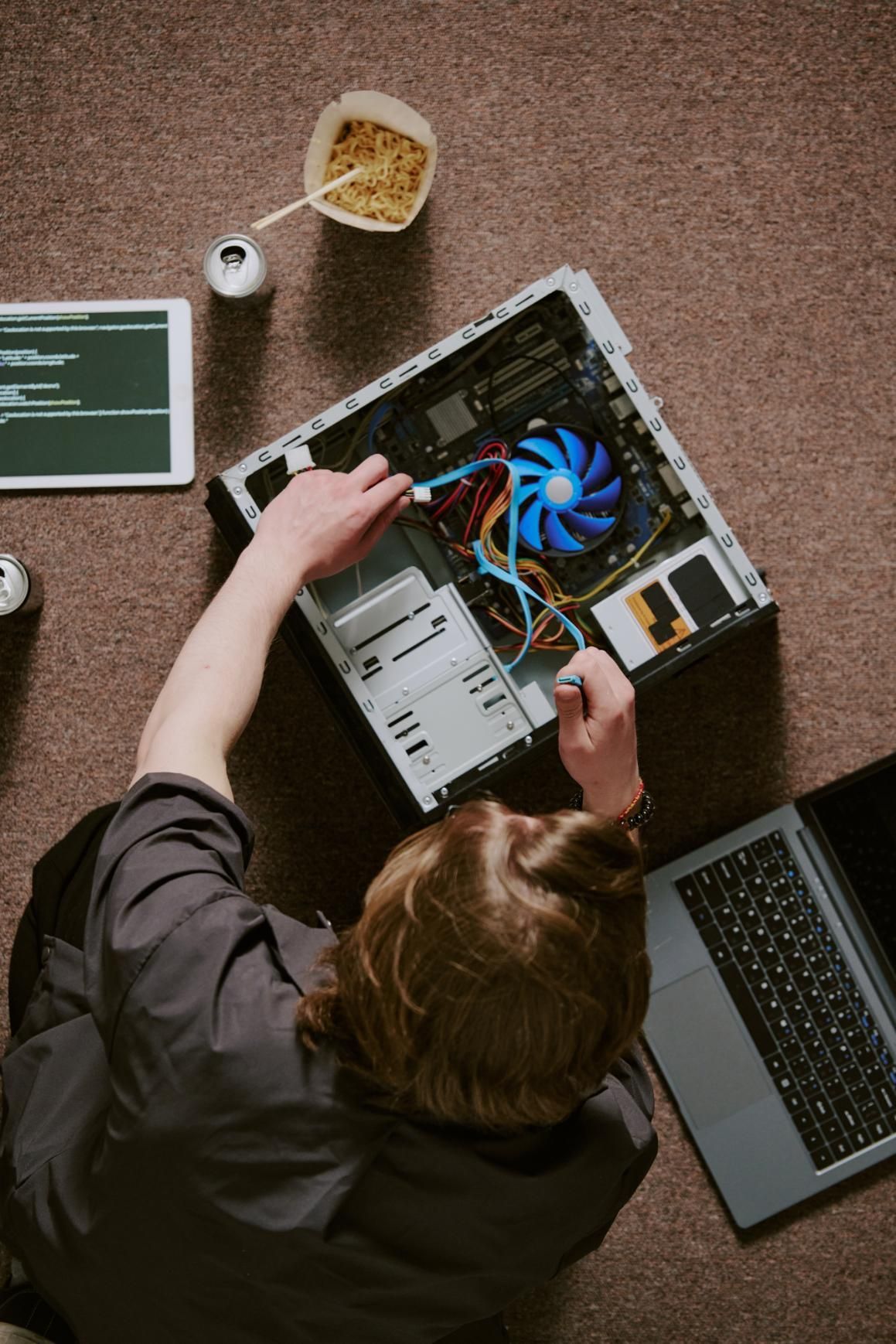Person working on a computer tower with tools, laptop, tablet, and snacks on the floor.