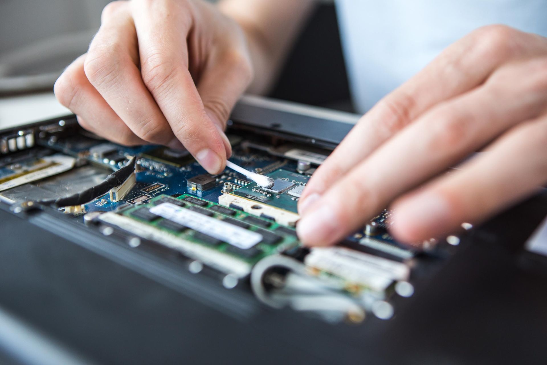 Hands cleaning a laptop's circuit board with a cotton swab, close-up shot of the device's interior.