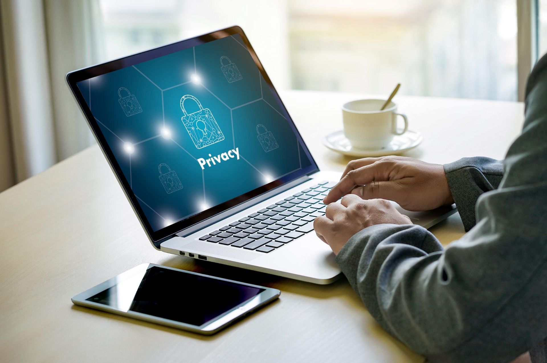 Person using a laptop with a privacy symbol on screen, tablet, and coffee cup on a desk.