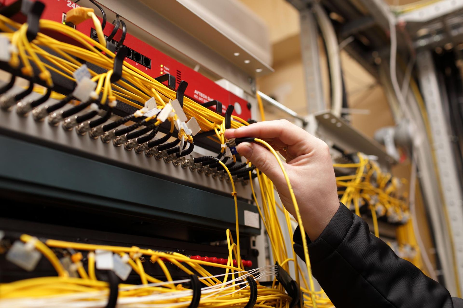 Person's hand connecting yellow fiber optic cables in a network server room with organized cables and equipment.