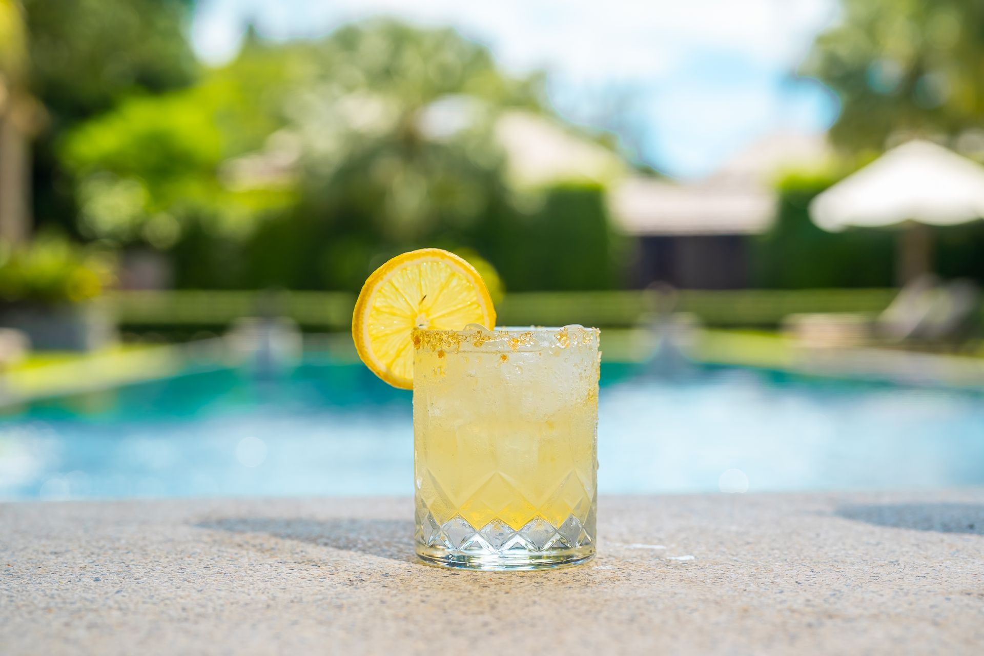 A glass of lemonade with a slice of lemon on a table next to a swimming pool.