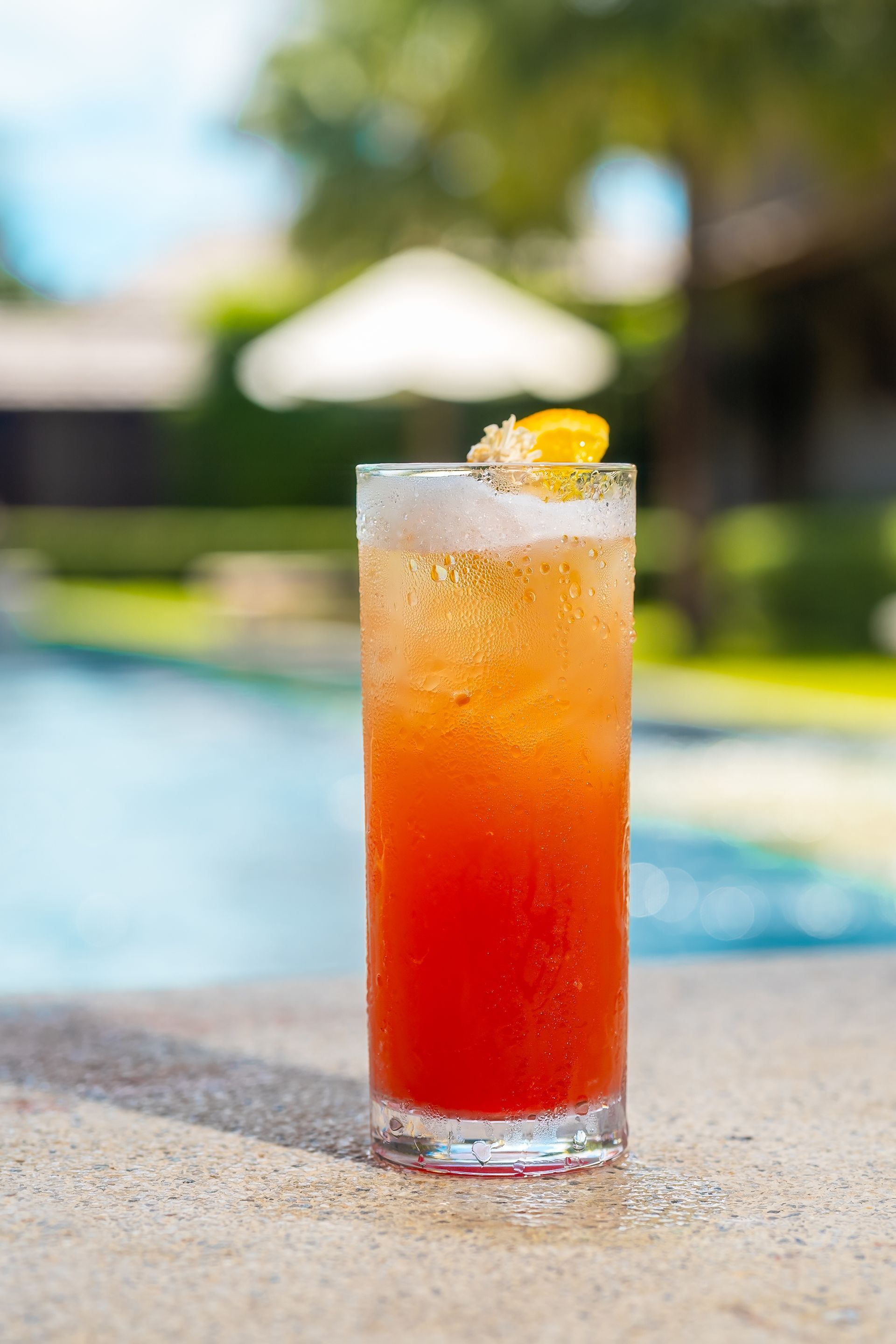 A tall glass filled with a red drink is sitting on the edge of a swimming pool.