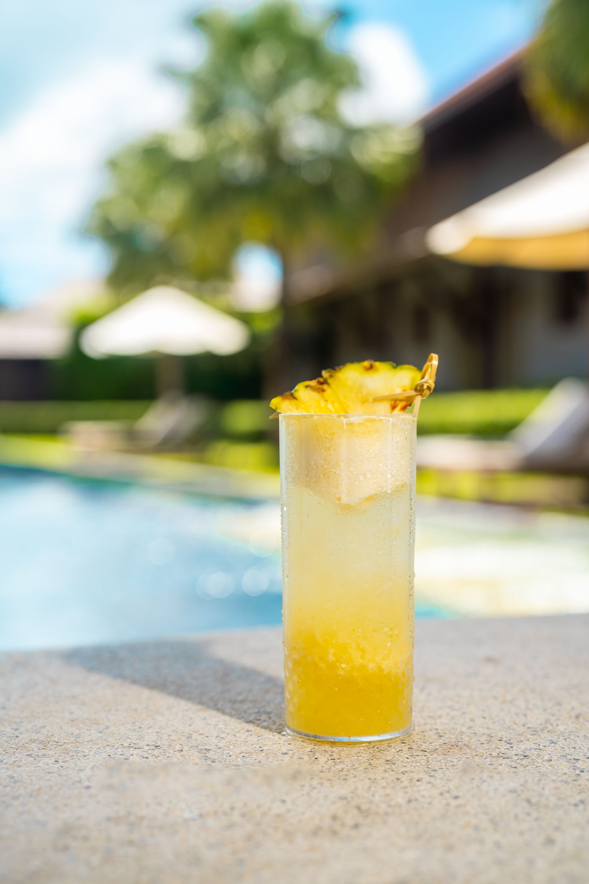 A pineapple cocktail in a tall glass on a table next to a swimming pool.