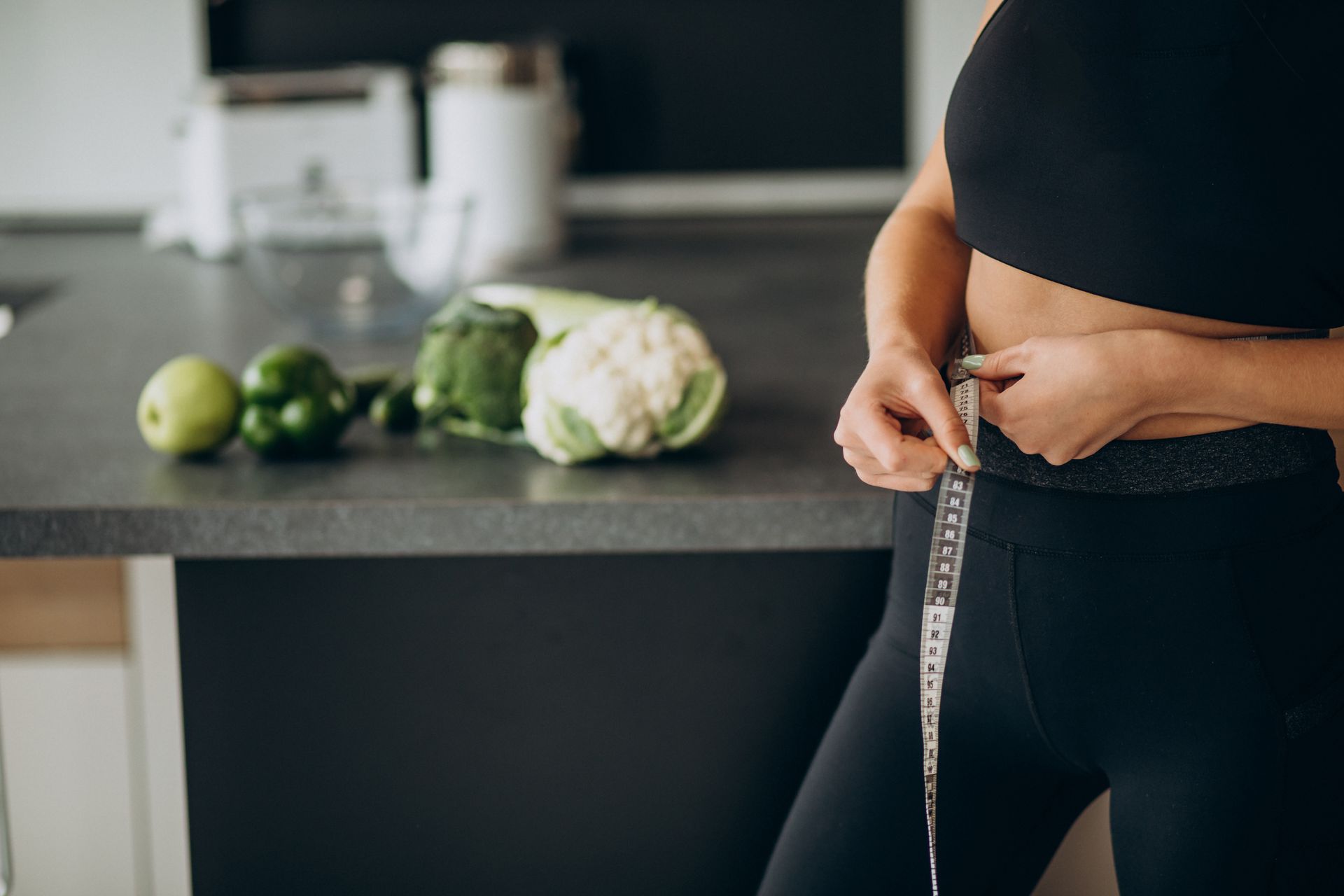 A woman is measuring her waist with a tape measure in a kitchen.