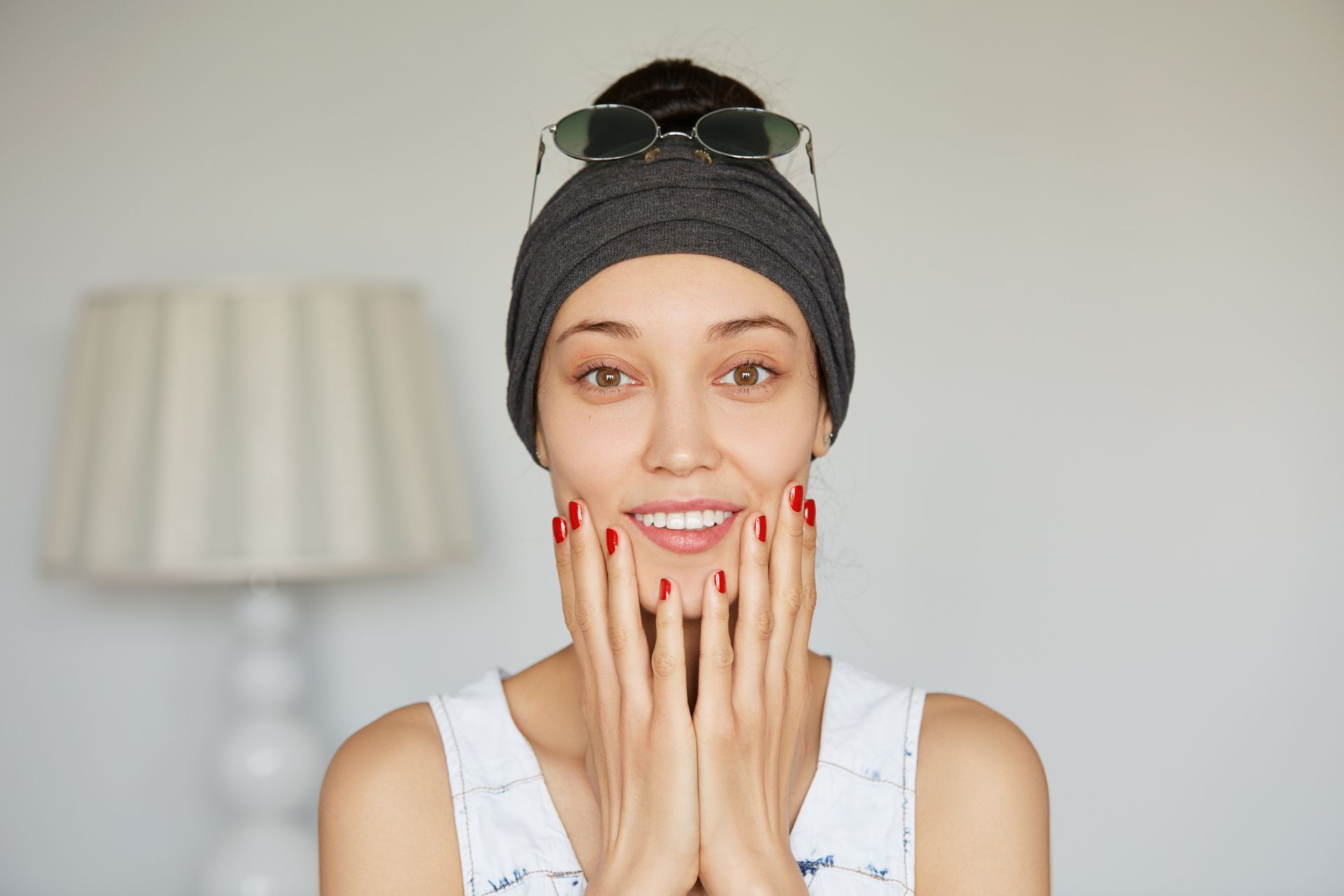 A woman wearing a headband and sunglasses is smiling with her hands on her face.