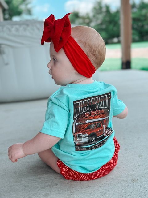 Baby wearing a red bow, turquoise shirt with a graphic, and red polka dot shorts, sitting outside.