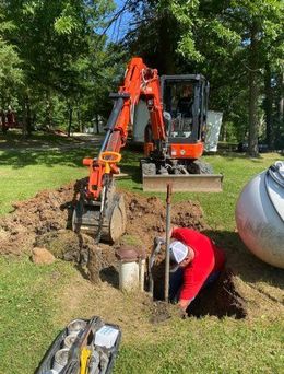Construction worker in trench, using a pole, with soil and pipes visible. Outdoor setting.