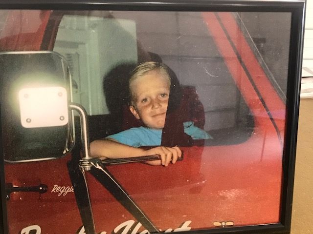 Boy in a red truck, smiling and leaning on the door. Window and mirror visible.