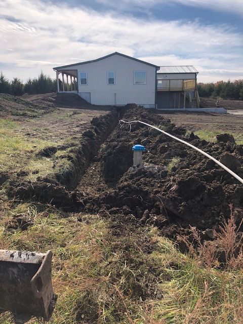 A trench dug in a field leads to a well near a white house with a porch.