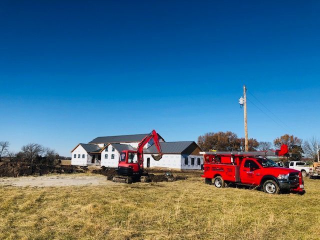Construction site: Red excavator digging near a new house under a clear, blue sky. Red service truck parked.