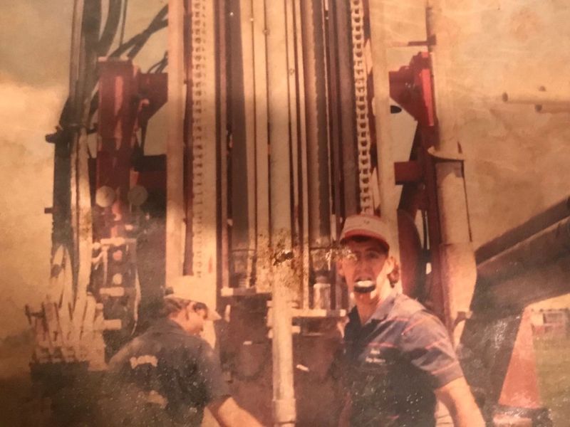 Two men working near a red and white drilling rig. One man smiles, holding a tool.