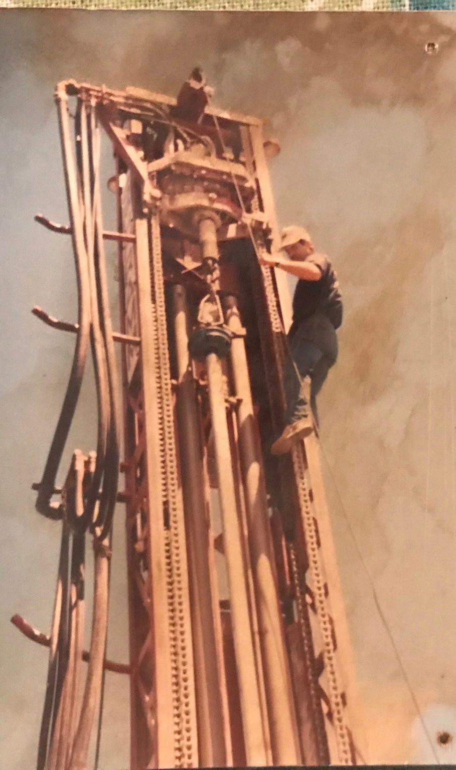 Man on drilling rig, inspecting machinery. Brown and gray metal, blue jeans, sunny outdoor setting.
