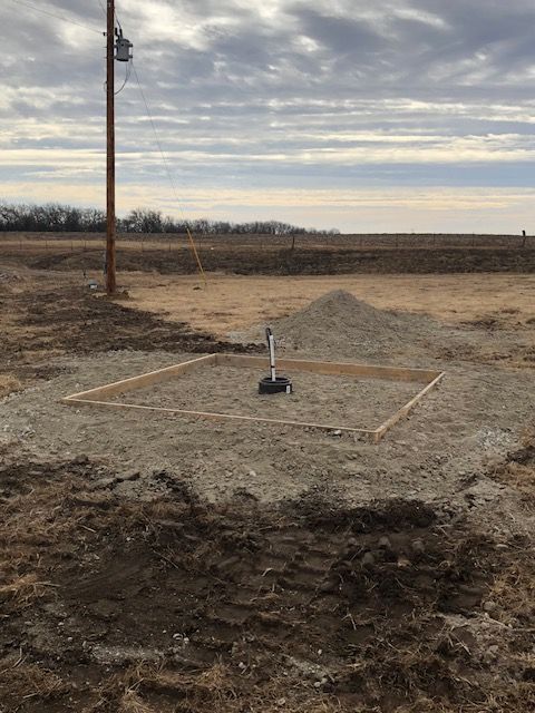 Well pipe with gravel surround and wooden frame, next to a utility pole in a field.