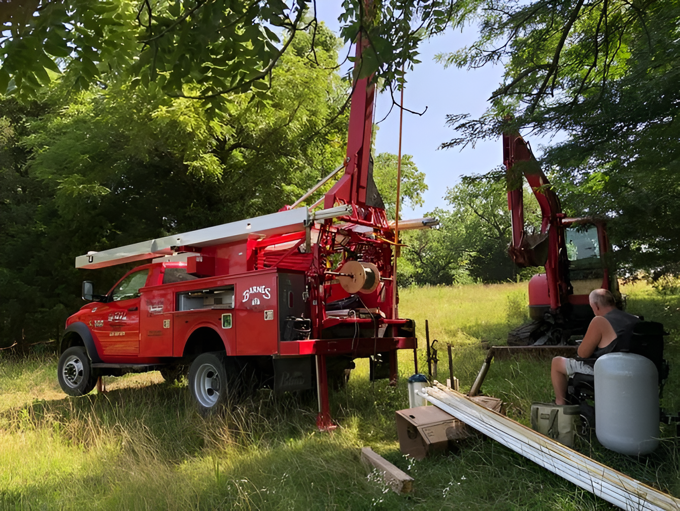 Red drilling truck and excavator in a grassy field, man sitting nearby, working on well.