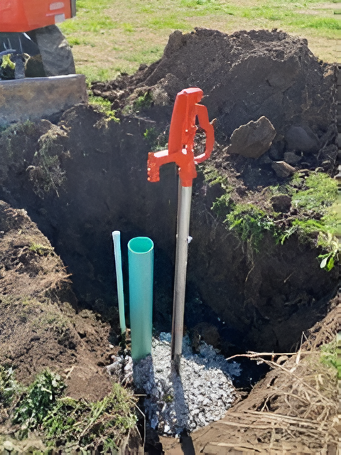 Orange hand pump, green and white pipes in a trench. Gravel base. Dirt pile in the background.