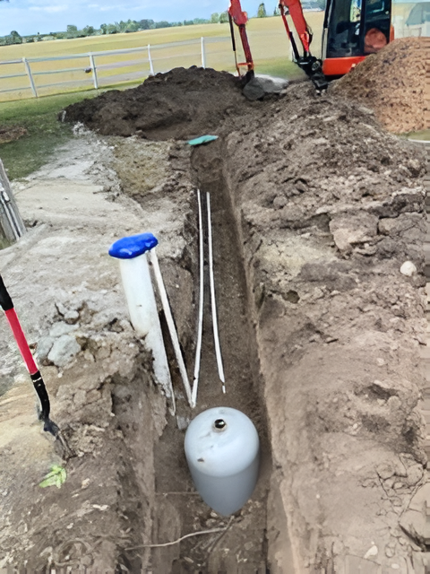 Trench with propane tank and piping, next to white fence. Backhoe in background.