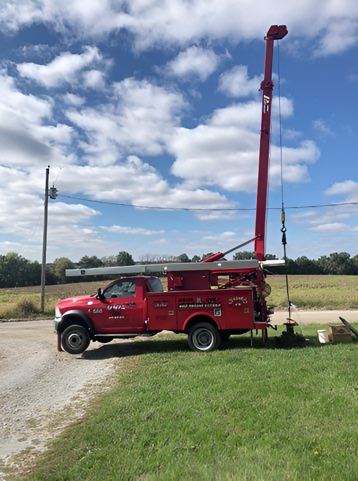 Red truck with drilling equipment on a grassy area next to a field under a cloudy blue sky.