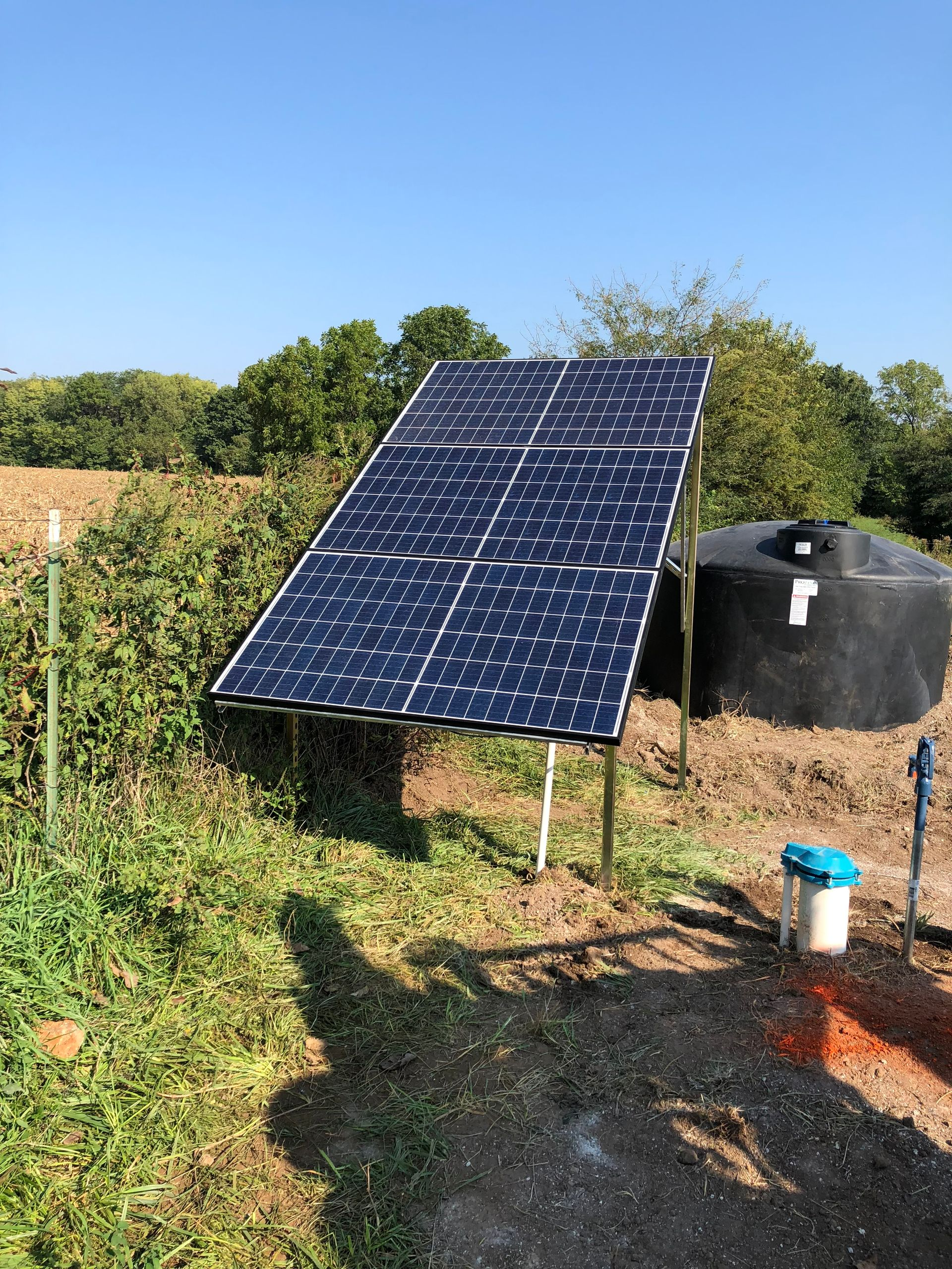 Solar panel tilted towards the sun, powering a water tank in a field.