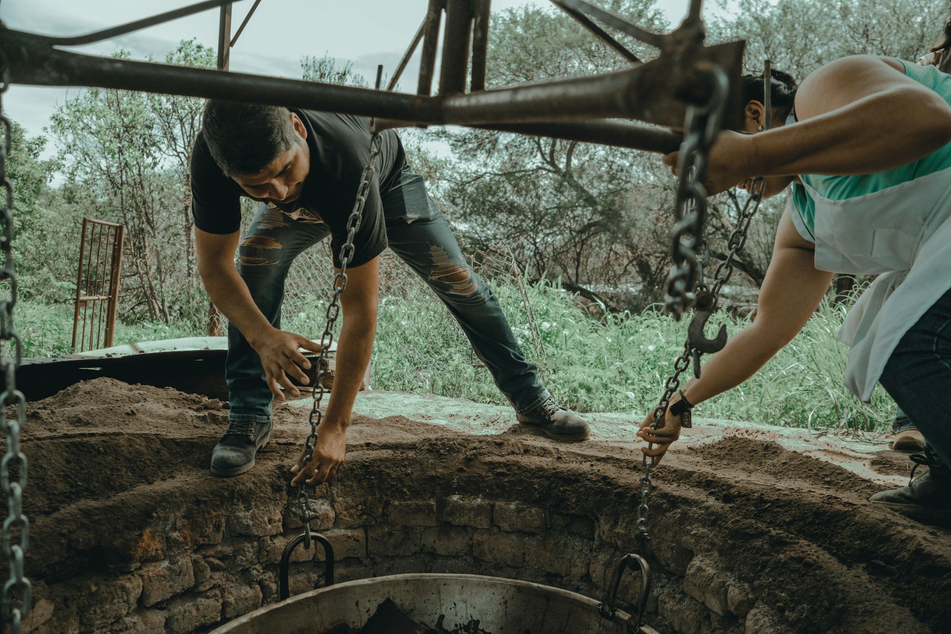 Two people lowering chains into a dark, earthen pit; outdoors.