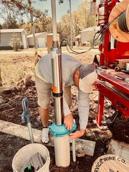 Man working on a large, blue pump outdoors. The man wears blue jeans, a hat, and work boots.