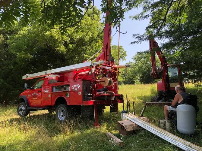 Red well drilling truck and excavator in a grassy field. A person sits near a white tank.