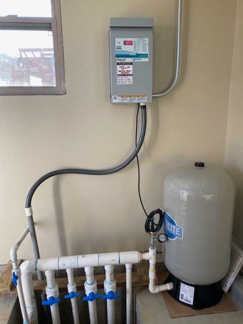 Electrical panel mounted above a water pressure tank and PVC piping with blue valves near a window.