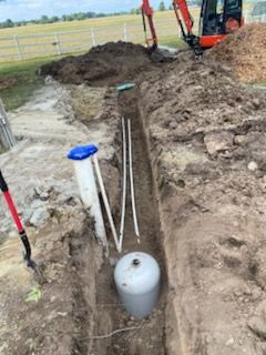 Trench with white pipes and a tank, next to dirt pile, small excavator, and a white fence in a field.