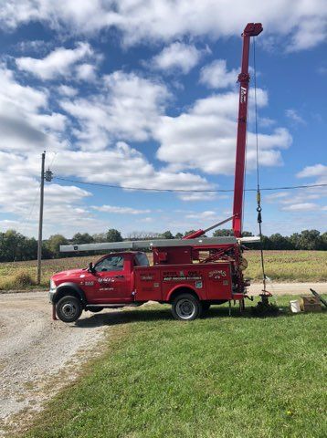 Red drilling rig mounted on a truck parked on a grassy area with blue sky and clouds.