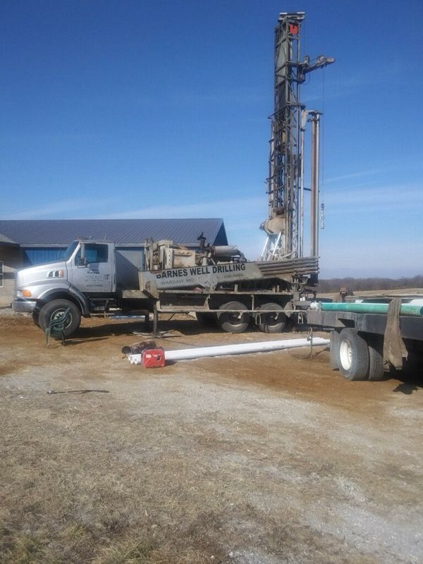 Drilling rig on a truck, outdoors on a sunny day. White pipe and trailer visible.