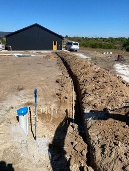 Trench dug in dirt, with propane tank, PVC pipe, and backhoe in background. Outdoor construction.