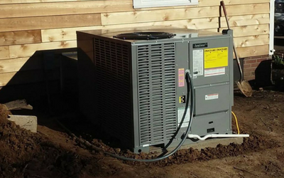 A gray HVAC unit sits on a concrete pad next to a wooden-sided house with dirt excavated around the base.