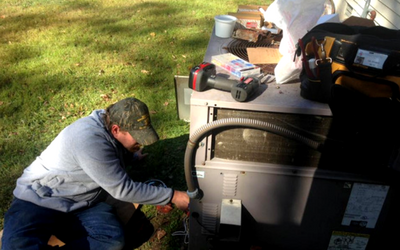 A person wearing a grey hoodie and camouflage cap repairs an outdoor HVAC unit on a sunny lawn.