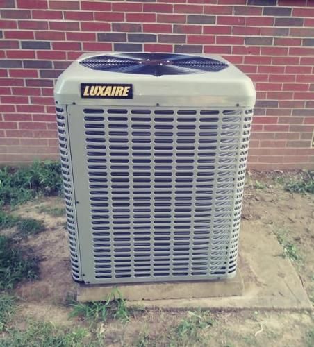A light gray Luxaire air conditioning unit sits outdoors on a concrete pad against a red brick wall.