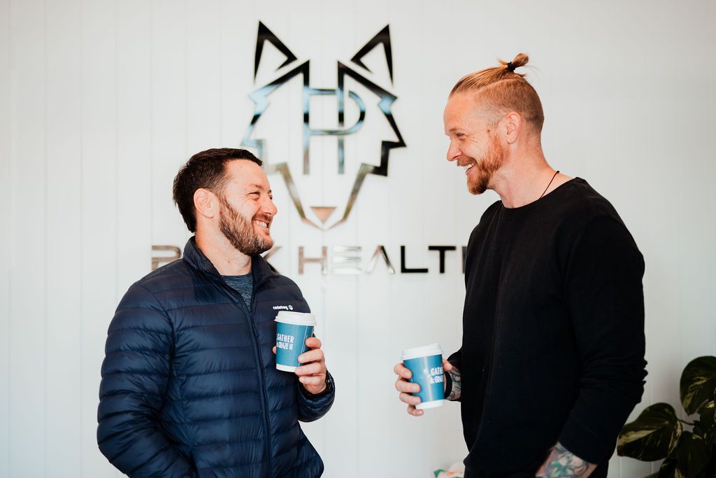 Two men are standing next to each other holding cups of coffee and having a laugh.