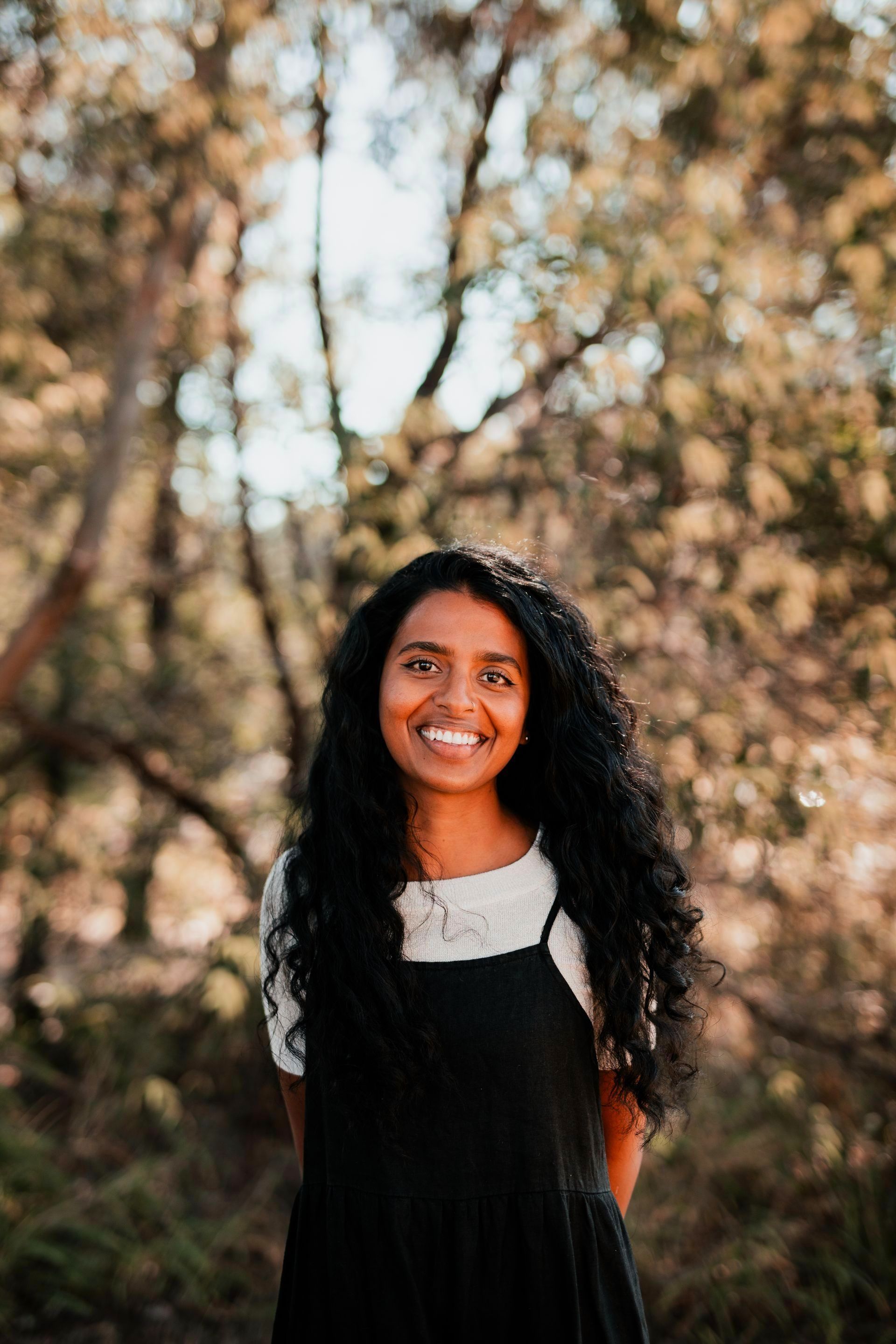 A woman in a black dress is smiling in front of a forest.