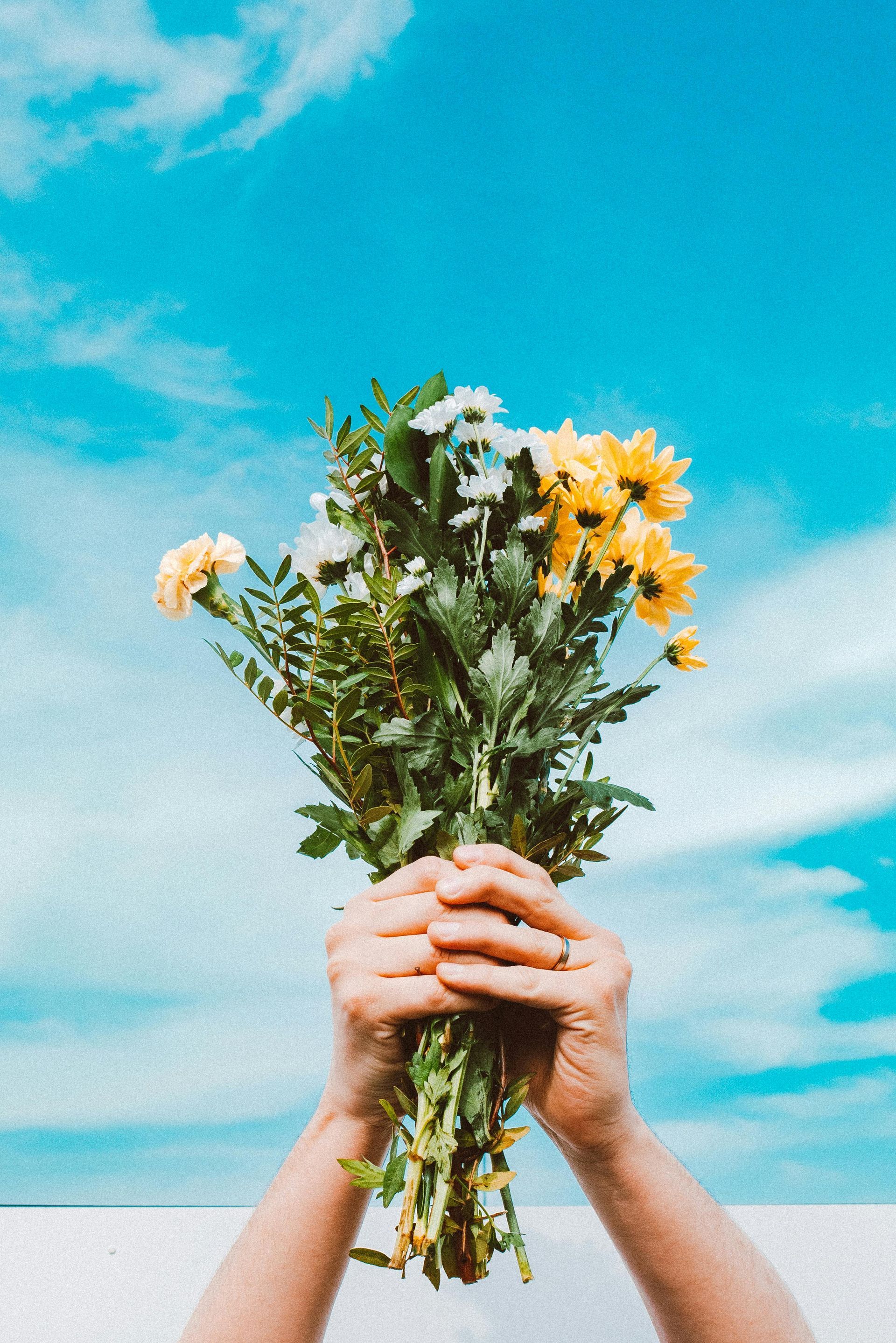 Hands holding a bouquet of yellow and white flowers against a bright blue sky.