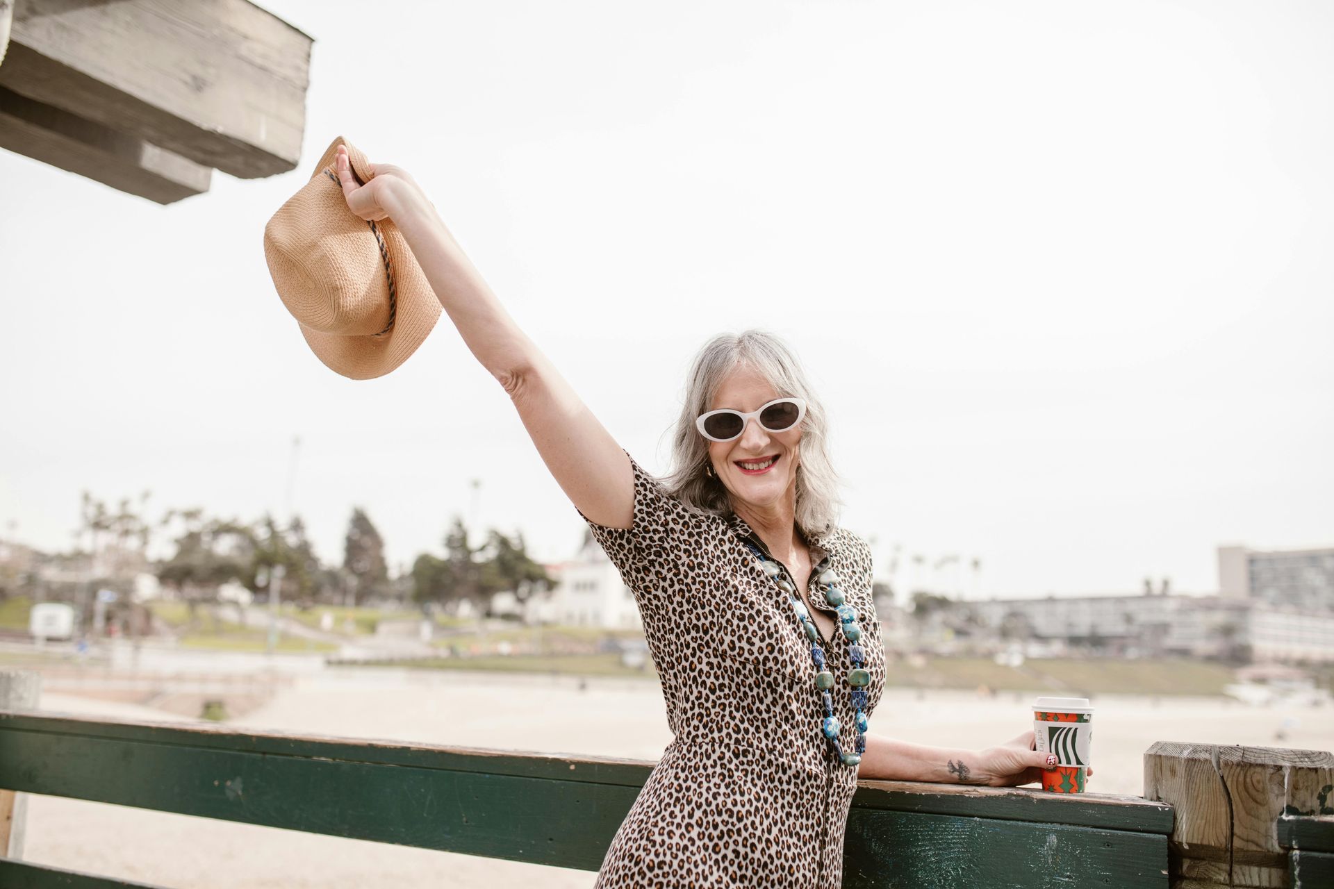 Woman with gray hair, sunglasses, and leopard print dress raises a hat at the beach, smiling.